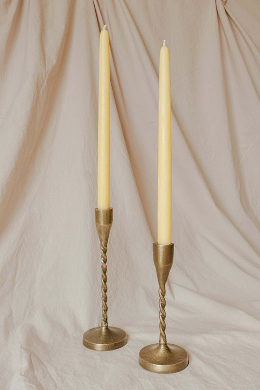 Two tall candles in gold candle holders on a beige fabric background
